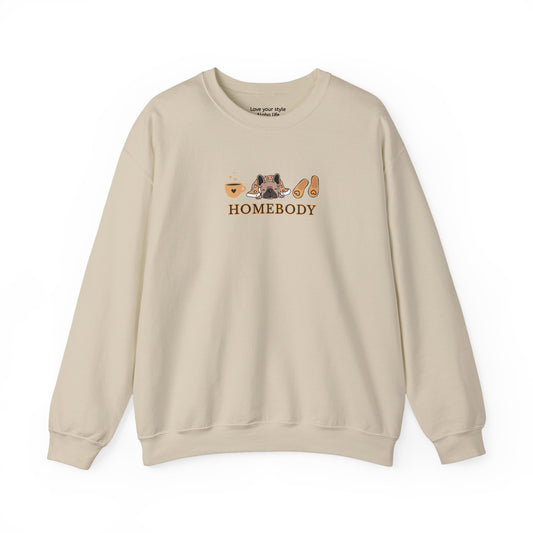 A beige crewneck sweatshirt featuring a central graphic of a dog under a blanket, house slippers, coffee cups and the word 'HOMEBODY' printed below it.