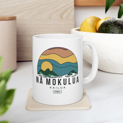 A white ceramic mug with a colorful retro-style landscape graphic and the text 'NA MOKULUA KAILUA HAWAII' sits on a light wood coaster in a kitchen setting.