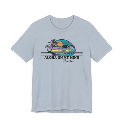 A light blue short-sleeve t-shirt with a graphic on the chest depicting a sunset over a beach wave, with the text 'ALOHA ON MY MIND' and 'Kailua' underneath.