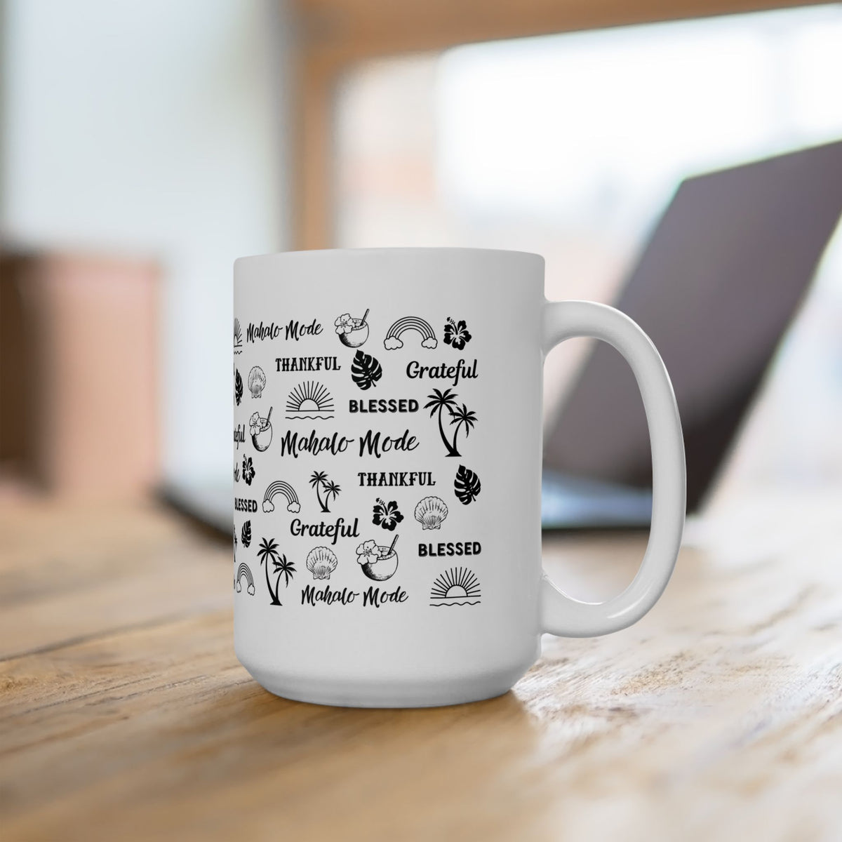 A white ceramic coffee mug with black printed Hawaiian and Mahalo-themed graphics and text, sitting on a wooden table with a blurred laptop in the background.