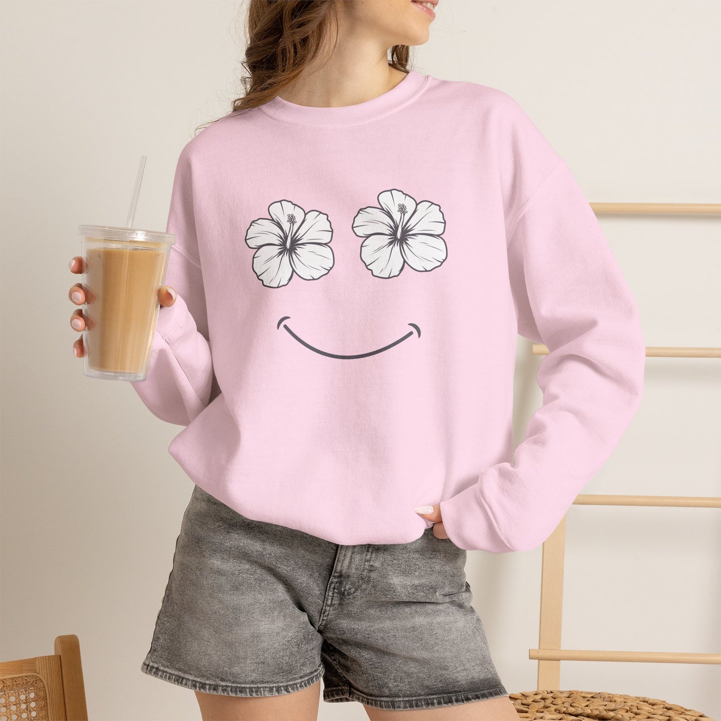 A woman wearing a light pink crewneck sweatshirt with a graphic of two white hibiscus flowers and a smiley face, paired with gray denim shorts and holding an iced coffee.