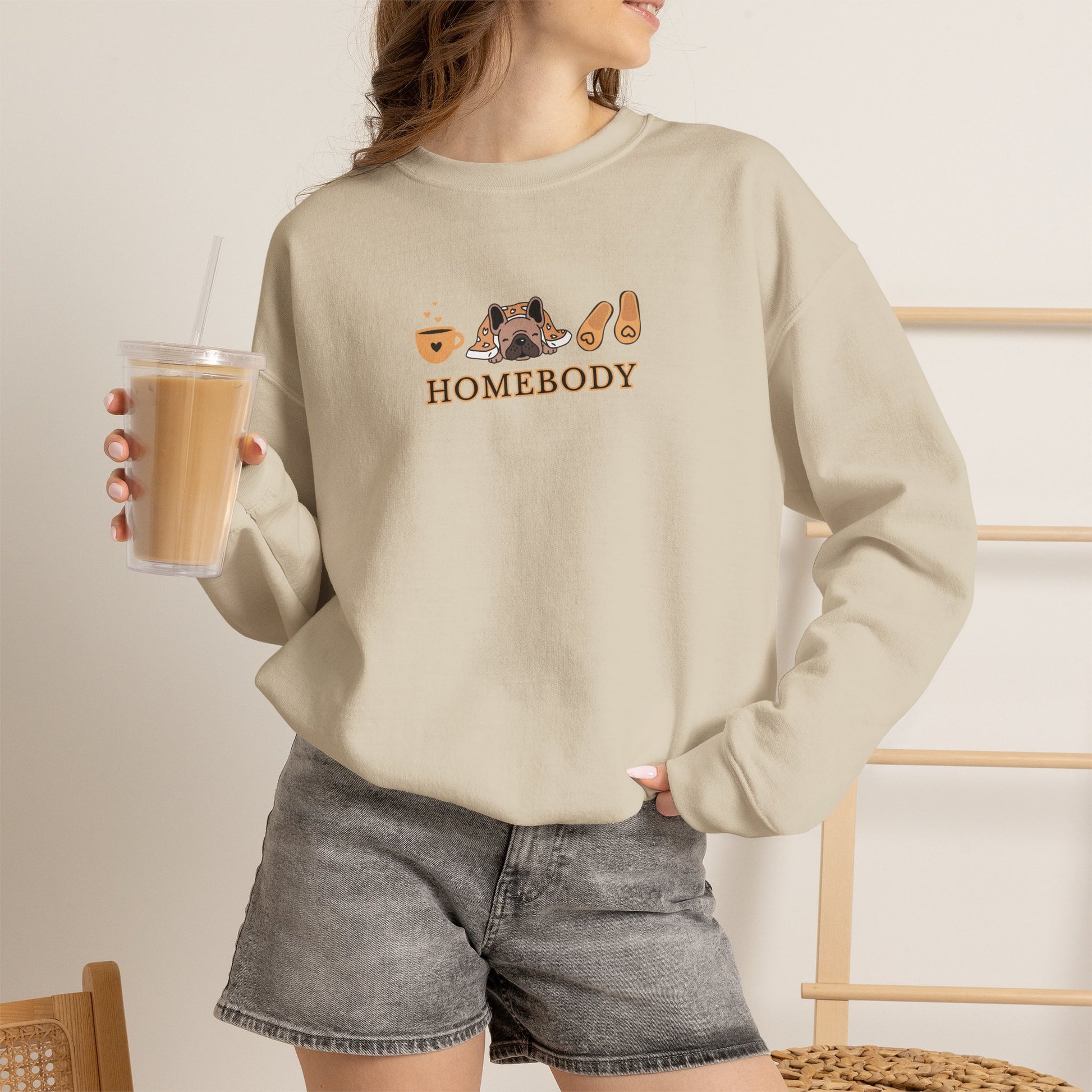 Person holding an iced coffee standing wearing a sand color crewneck sweatshirt featuring a central graphic of a dog under a blanket, house slippers, coffee cups and the word 'HOMEBODY' printed below it.