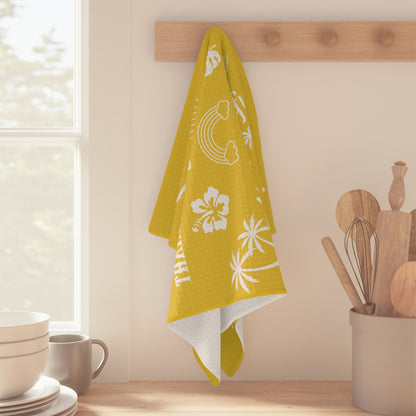 A yellow waffle-weave kitchen towel with white tropical-themed text 'mahalo mode' 'thankful' 'blessed' 'grateful' and illustrations, hanging from a clothesline with wooden clips against a white background hanging on a wood peg.