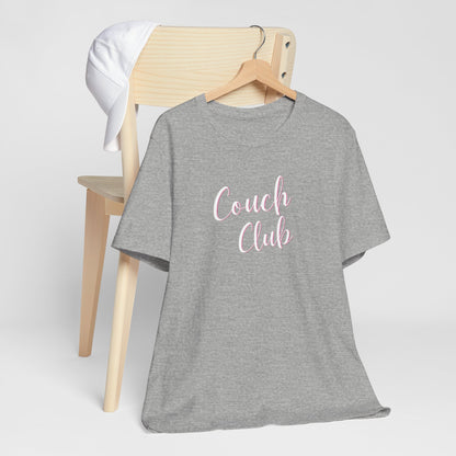 A heather gray short-sleeve t-shirt on a wooden hanger, styled in front of a light wood chair with a white cap. The t-shirt features the text 'Couch Club' in white script.