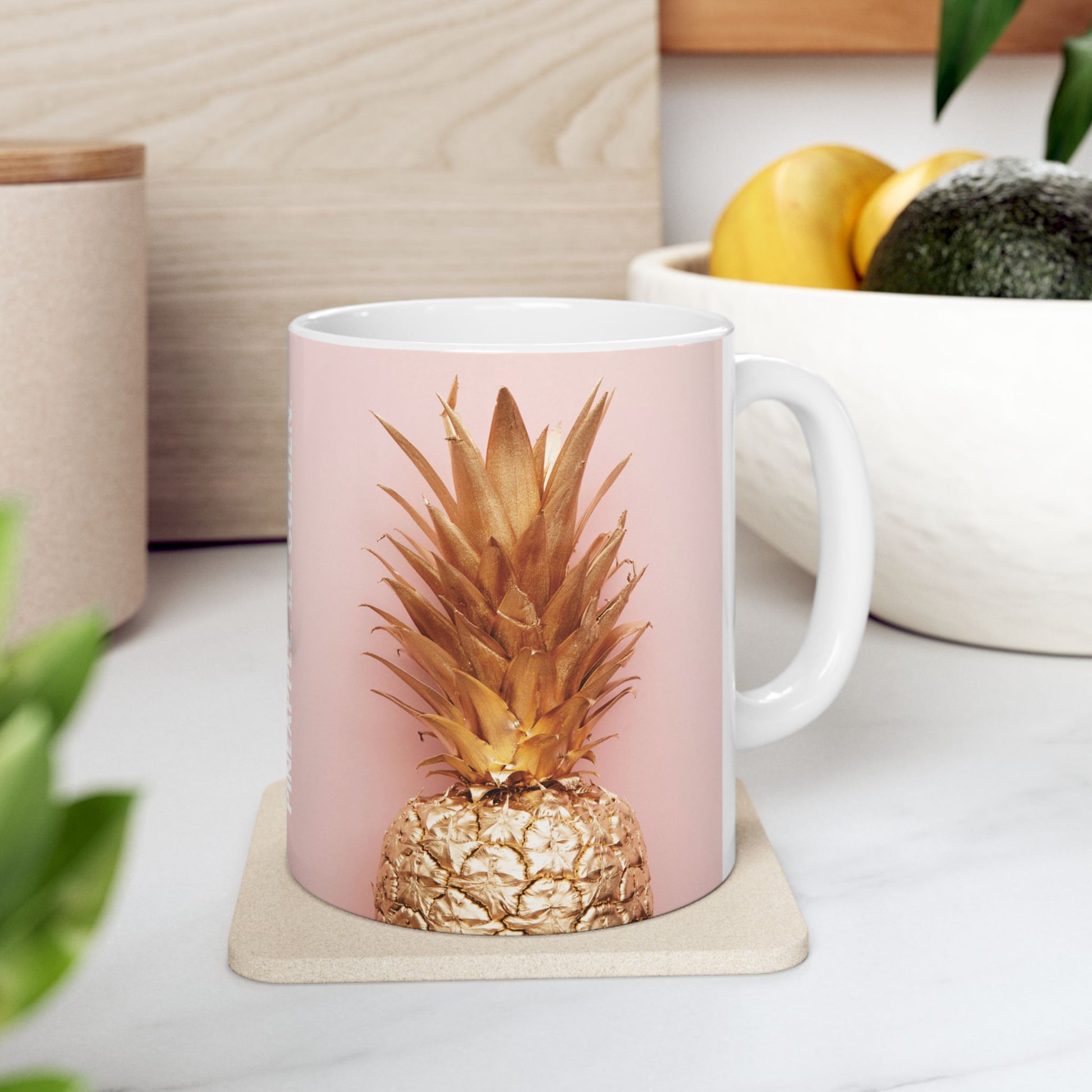A white ceramic mug with a pale pink printed background and a metallic gold pineapple design, sitting on a beige coaster on a kitchen counter.