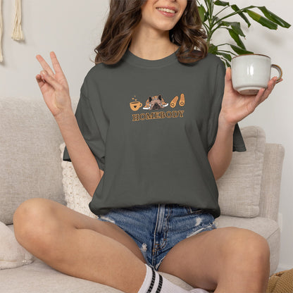 A woman sitting on a light beige couch wearing a dark gray t-shirt with a graphic that says 'HOMEBODY' and shows a mug in one hand and peace sign in the other.