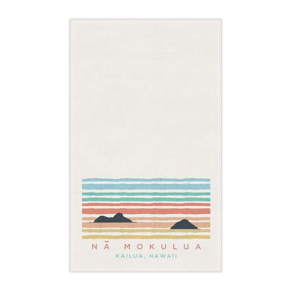 An off-white rectangular towel featuring a retro-style sunset illustration with horizontal stripes in blue, green, coral, and yellow, along with two dark mountain silhouettes and the text 'NĂ MOKULUA KAILUA, HAWAII' at the bottom.