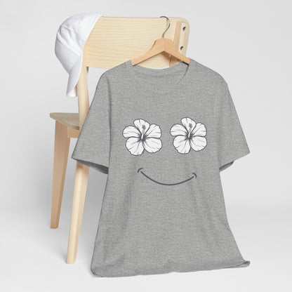 A heather gray short-sleeve t-shirt on a wooden hanger, featuring a graphic of two white hibiscus flowers for eyes and a curved smile. The shirt is styled on a light wood chair with a white towel draped over the backrest, against a clean light gray background.