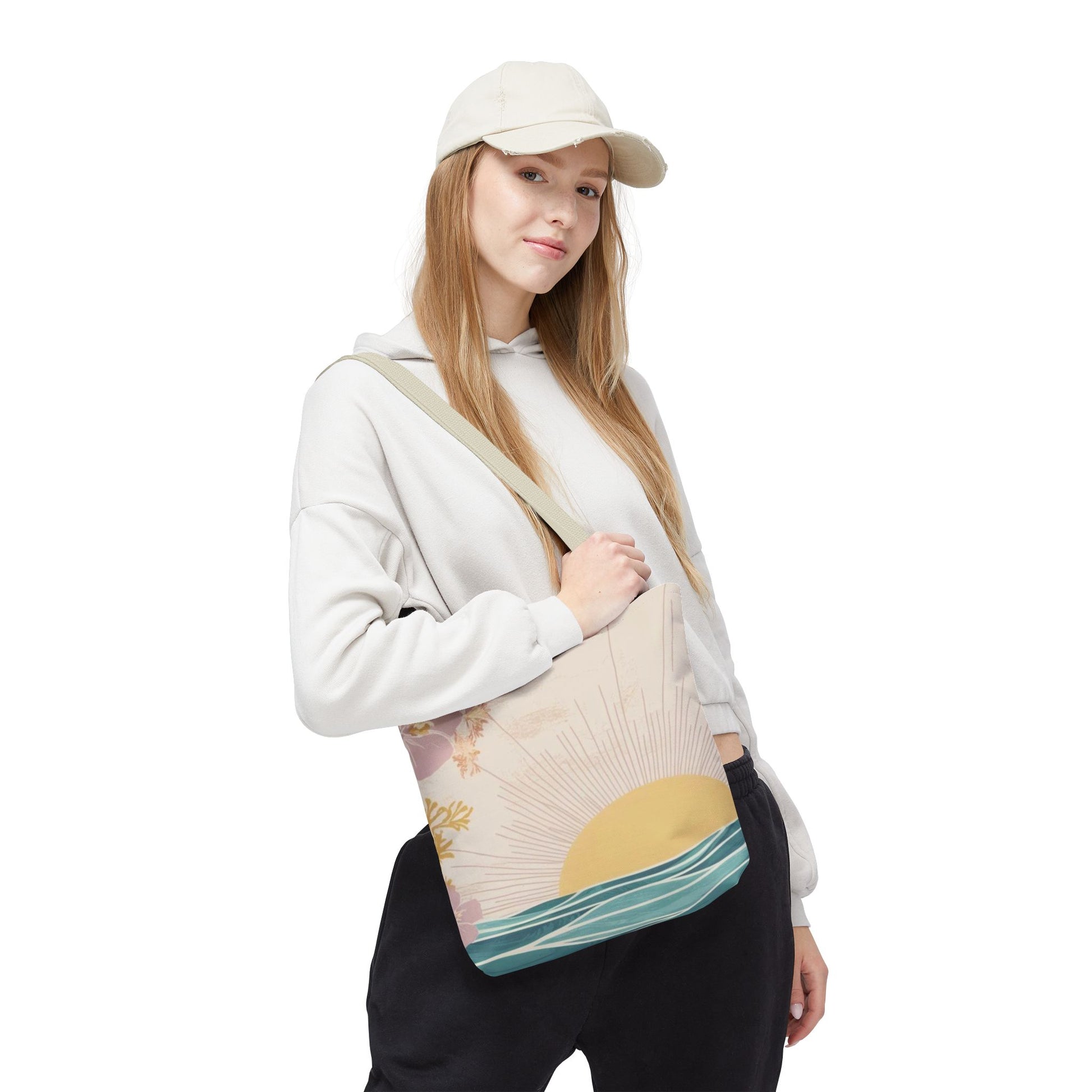 A woman wearing a cream baseball cap, white hoodie, and black sweatpants carries a canvas tote bag with a sunset and ocean wave design. The bag has a beige background with a yellow sun, gold stars, and teal-blue waves.