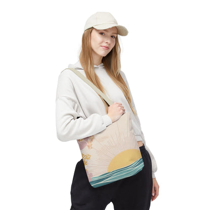 A woman wearing a cream baseball cap, white hoodie, and black sweatpants carries a canvas tote bag with a sunset and ocean wave design. The bag has a beige background with a yellow sun, gold stars, and teal-blue waves.