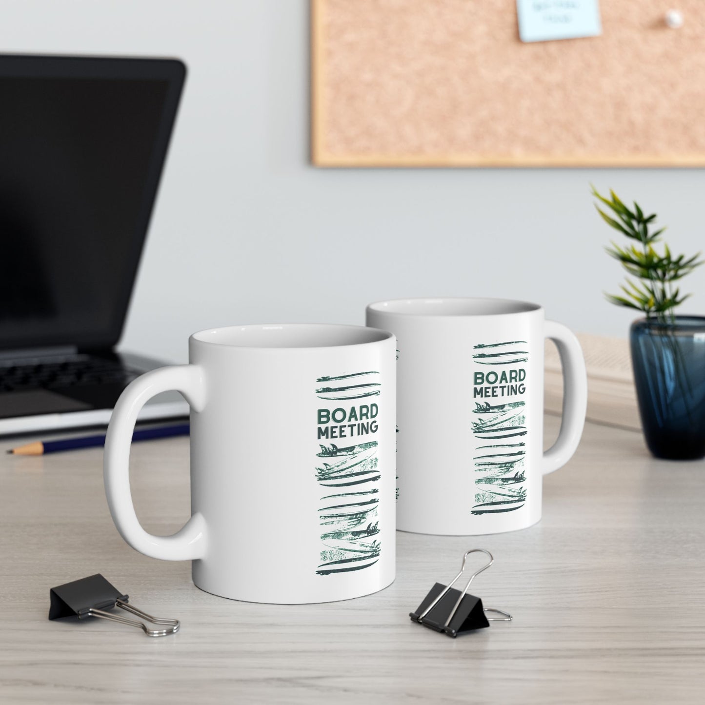 Two mugs with ʻBoard Meetingʻ illustrations and text on a desk with office items.