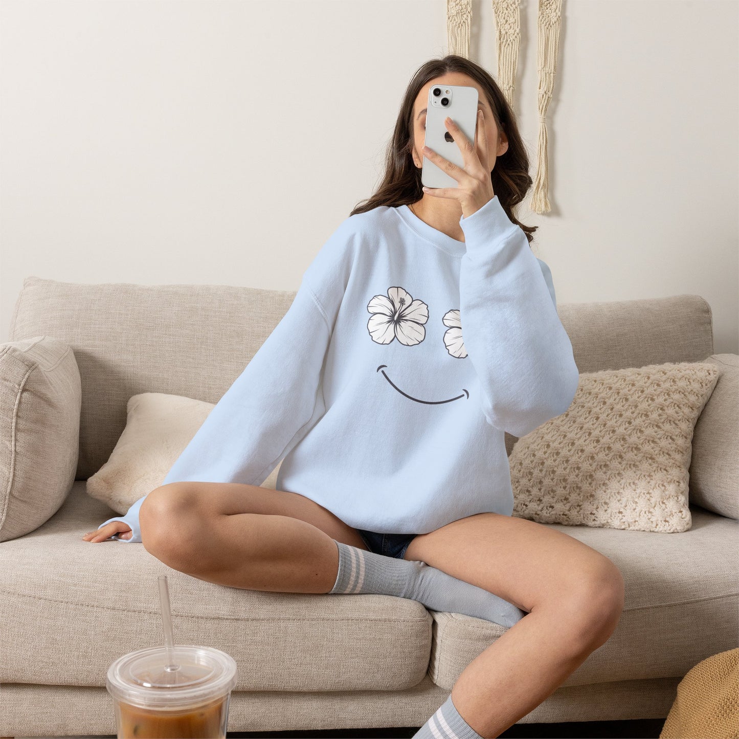 A woman sitting on a beige couch takes a mirror selfie while wearing a light blue sweatshirt with a white flower and black smiley face graphic. She is wearing gray socks and holding a phone, with an iced coffee in the foreground.