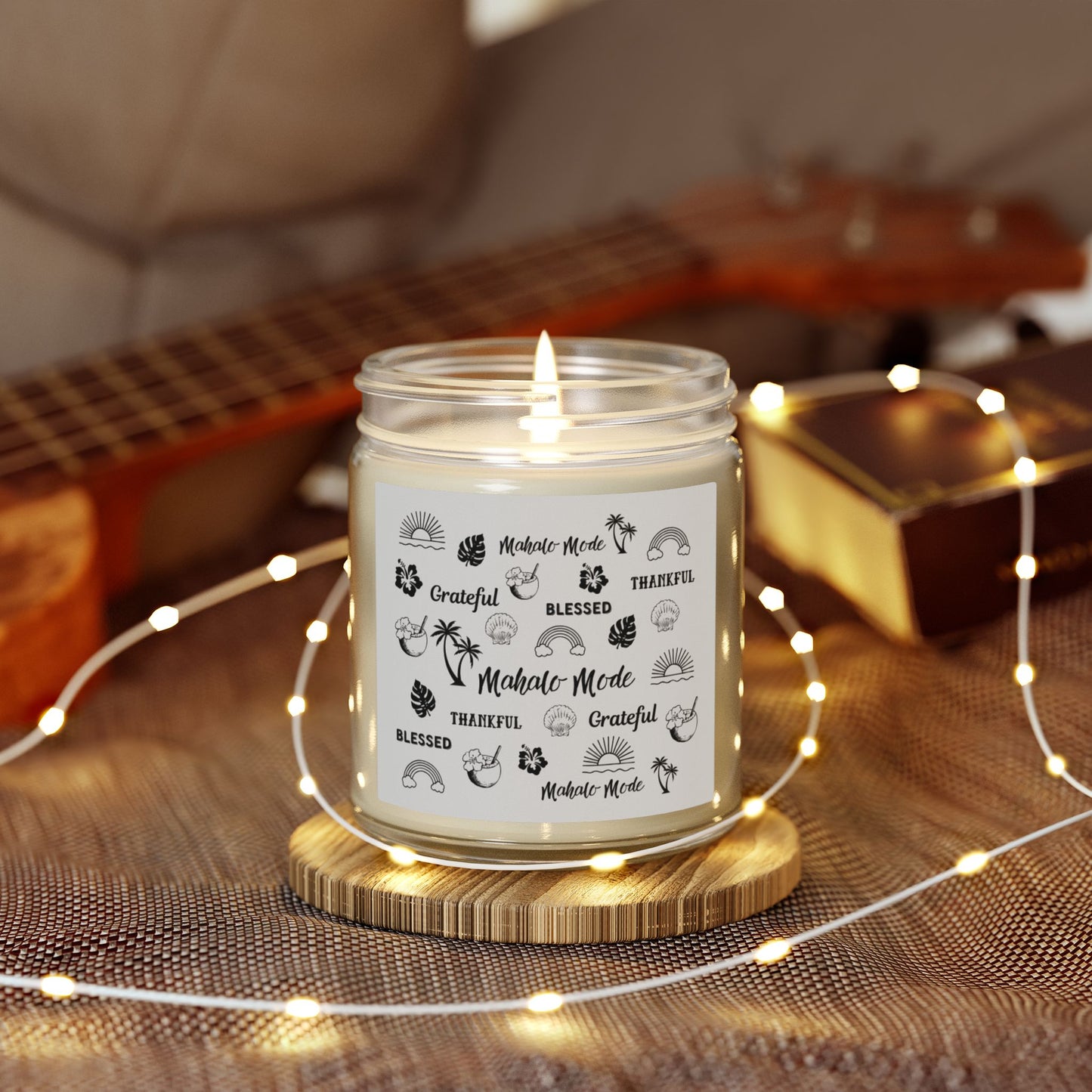 A lit white candle in a clear glass jar sits on a round wooden coaster, surrounded by warm fairy lights. The label features Hawaii tropical illustrations and the words 'Mahalo Mode', 'BLESSED', 'Grateful' and 'Thankful'