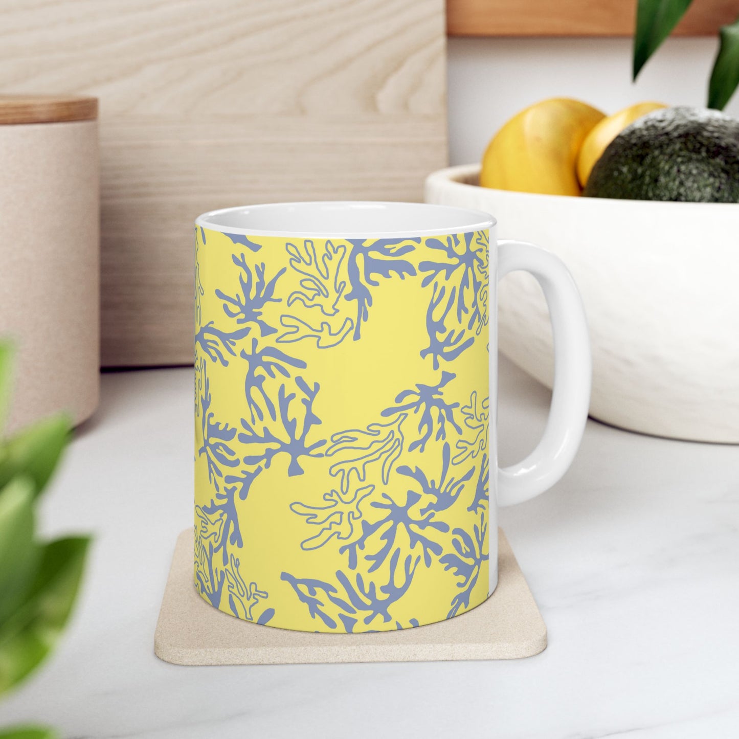 A white ceramic mug with a yellow and blue coral pattern, sitting on a light beige coaster on a white marble countertop in a kitchen setting.
