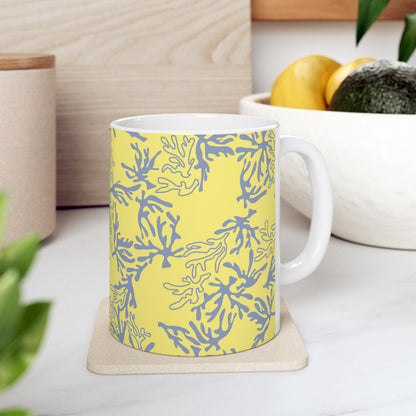 A white ceramic mug with a yellow and blue coral pattern, sitting on a light beige coaster on a white marble countertop in a kitchen setting.