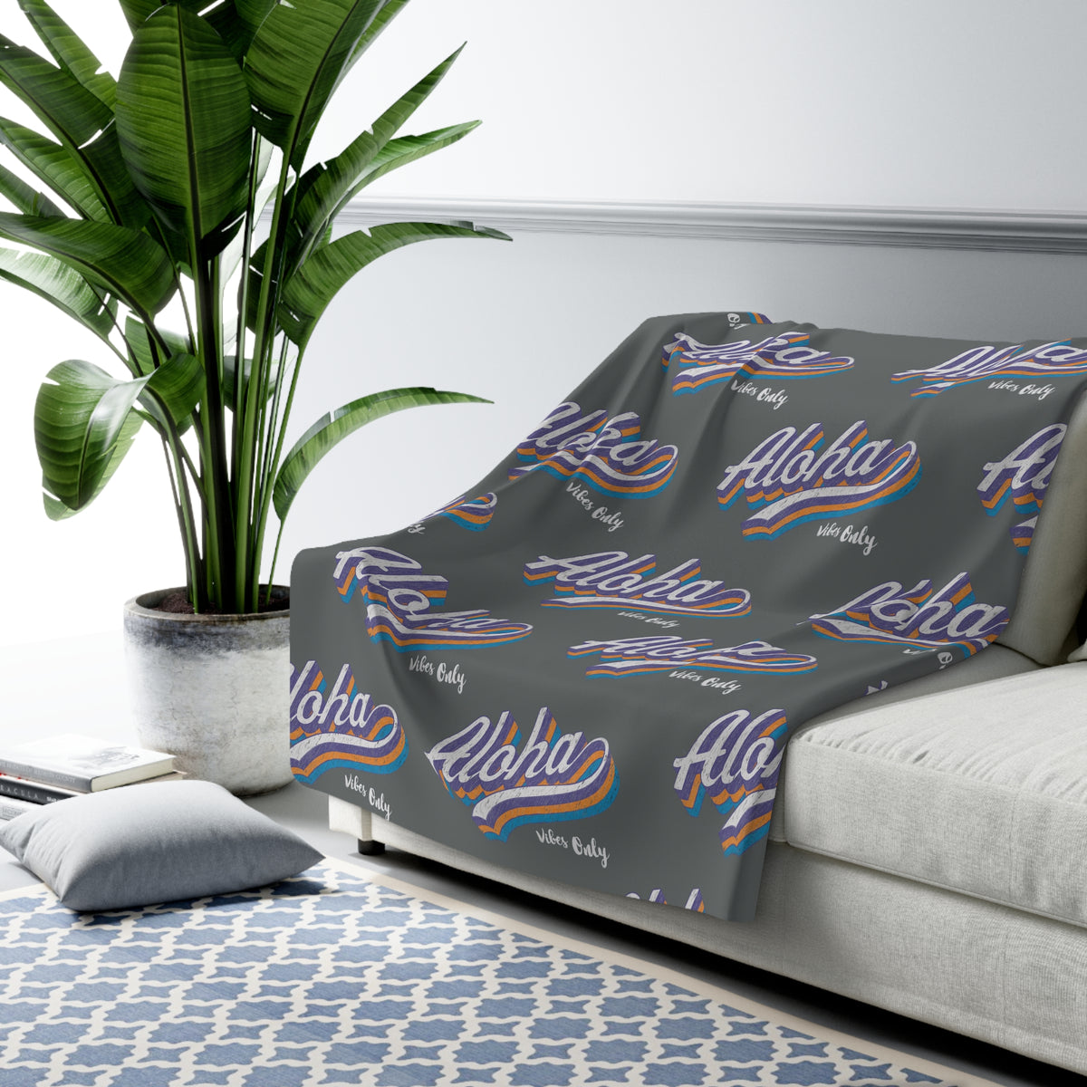 A gray throw blanket draped over a light gray sofa in a styled living room setting, featuring a repeating pattern of 'Aloha vibes only' in white script with blue and orange wavy stripes and 'Hawaii' in small white text.