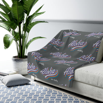 A gray throw blanket draped over a light gray sofa in a styled living room setting, featuring a repeating pattern of 'Aloha vibes only' in white script with blue and orange wavy stripes and 'Hawaii' in small white text.