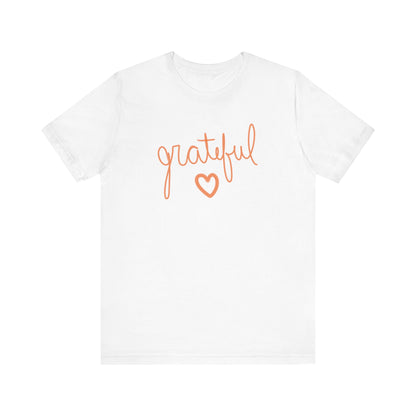 A white short-sleeve crewneck t-shirt featuring the word 'grateful' in orange cursive script and a matching orange heart outline on the chest, displayed against a solid white background.