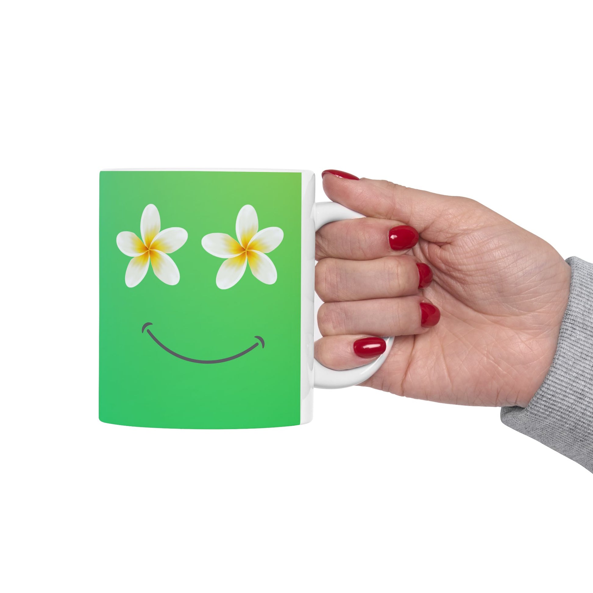 A hand with red nail polish holds a white ceramic mug against a plain white background. The mug features a green background with two white plumeria flowers for eyes and a curved smile.
