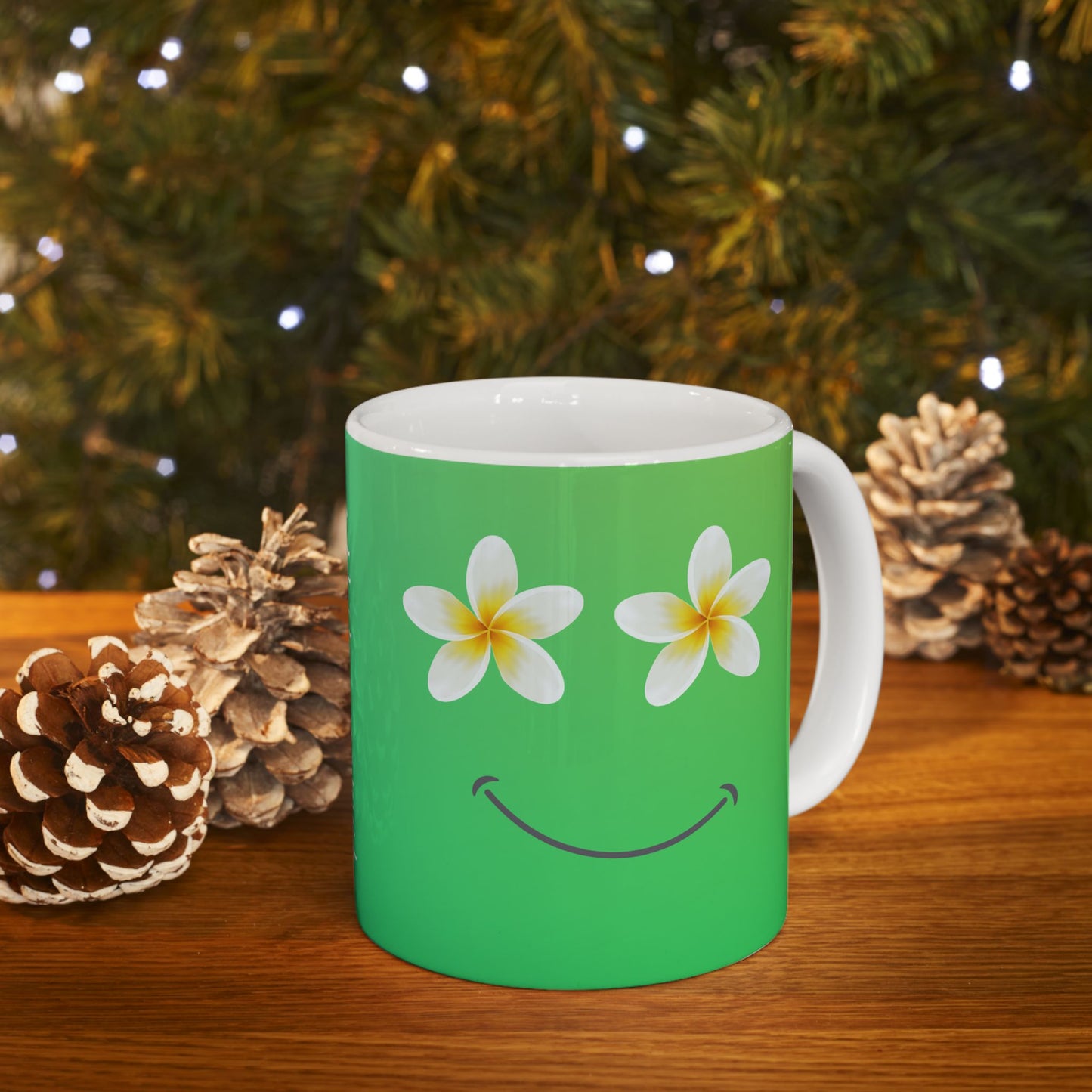 A green ceramic mug with a white handle and interior, decorated with two white plumeria flowers for eyes and a curved smile, set on a wooden surface with pinecones and blurred Christmas tree lights in the background.
