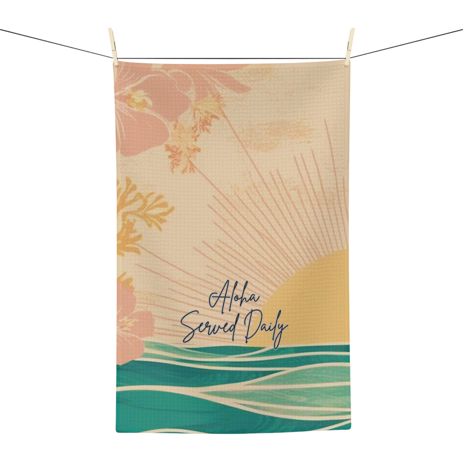 A rectangular kitchen towel with a tropical beach illustration hangs from a string with two wooden clips. The towel features a beige background with a yellow sunburst, pink hibiscus flowers, teal ocean waves, and the text 'Aloha Served Daily' in navy script.