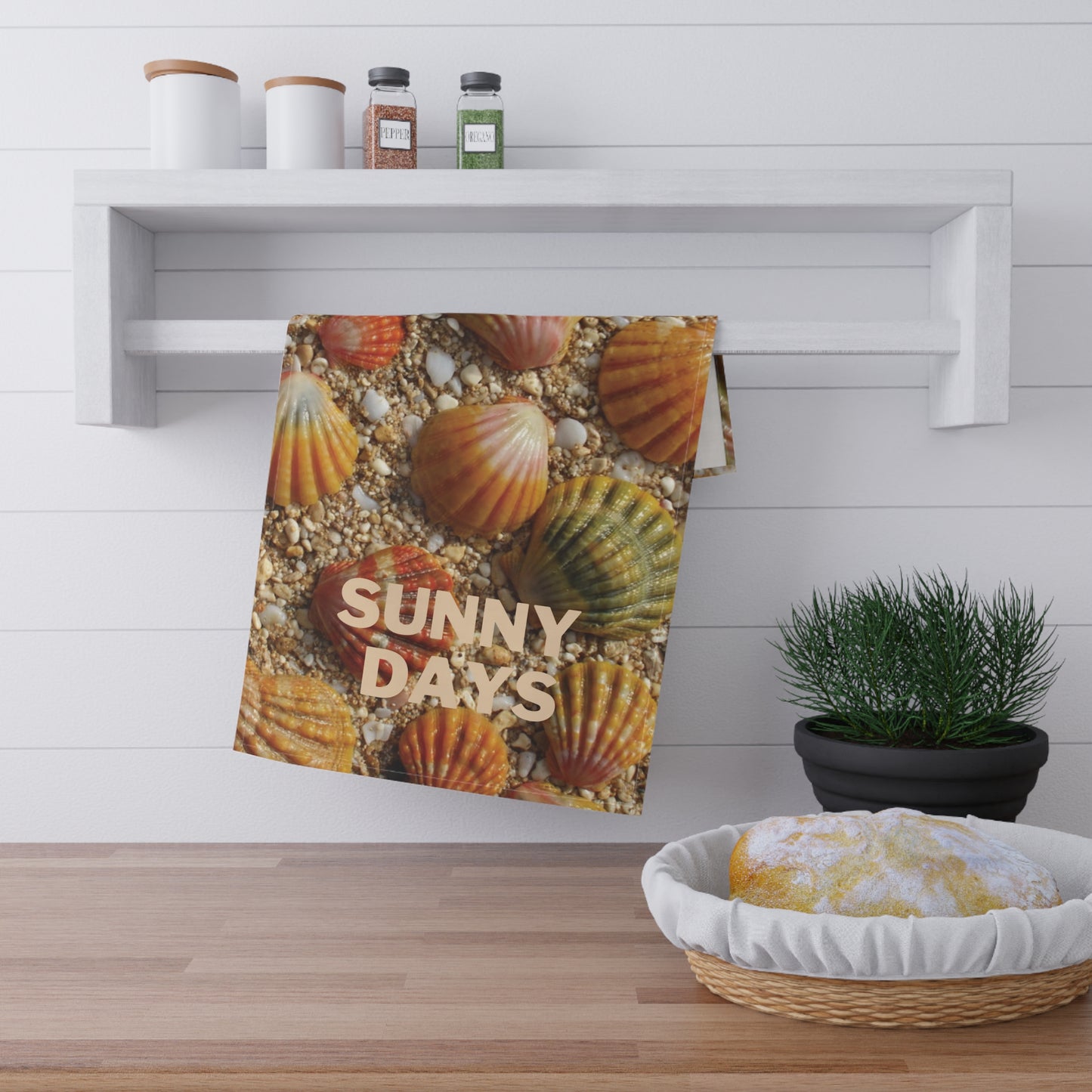 A kitchen towel featuring a colorful print of sunrise seashells on sand with the text 'SUNNY DAYS' in beige, hanging from a white wall rack in a bright kitchen setting.
