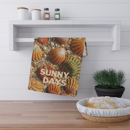 A kitchen towel featuring a colorful print of sunrise seashells on sand with the text 'SUNNY DAYS' in beige, hanging from a white wall rack in a bright kitchen setting.