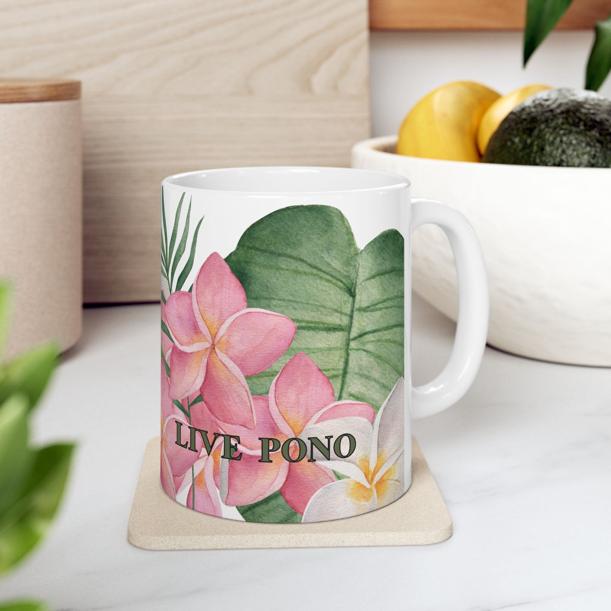 A white ceramic mug with a tropical floral print featuring pink plumeria flowers, green leaves, and the text 'LIVE PONO', sitting on a light wood coaster in a kitchen setting.
