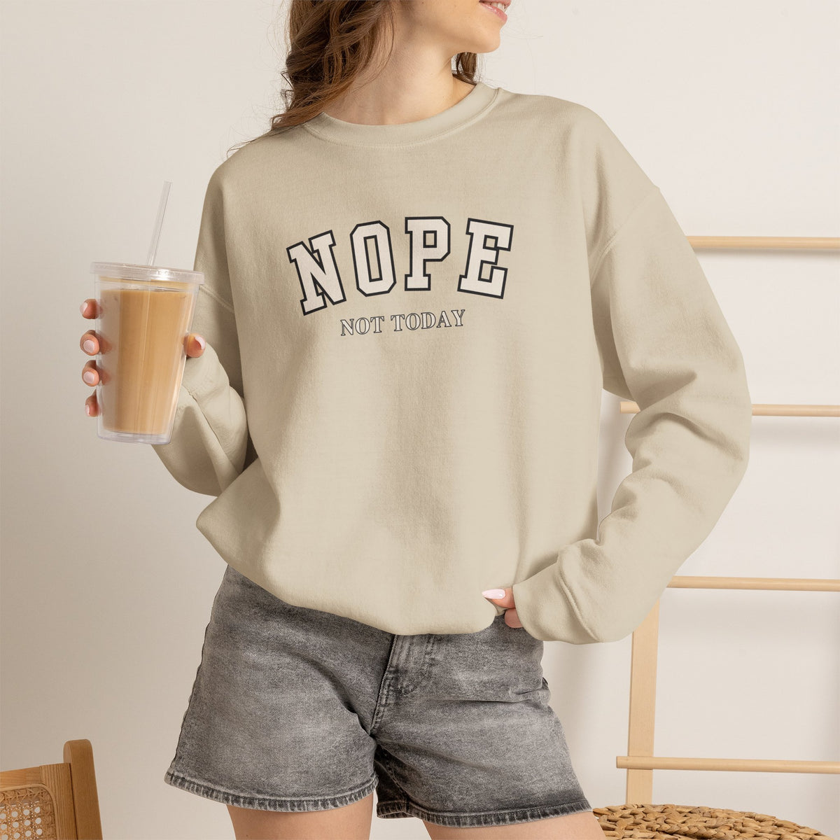 A woman wearing a SAND crewneck sweatshirt with black text reading 'NOPE NOT TODAY', paired with gray denim shorts, holding an iced coffee.