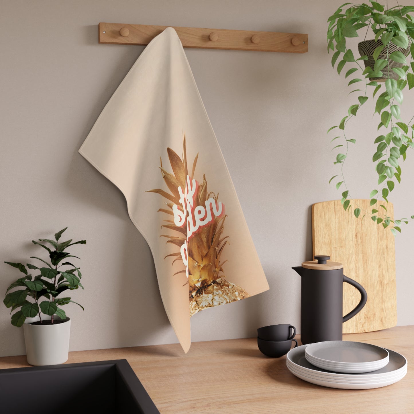 A beige kitchen towel with a graphic of a pineapple and the word 'stay golden' in pink script, hanging from a wooden peg on a kitchen wall above a countertop with dishes and plants.