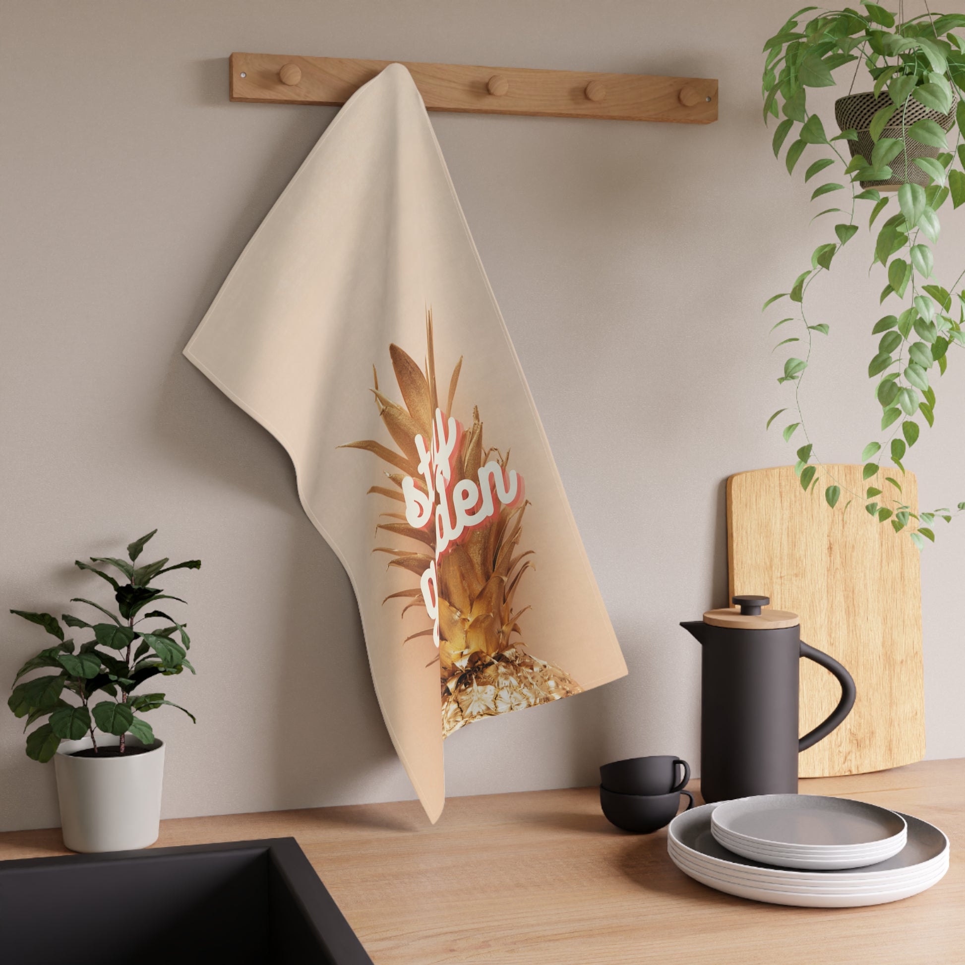 A beige kitchen towel with a graphic of a pineapple and the word 'stay golden' in pink script, hanging from a wooden peg on a kitchen wall above a countertop with dishes and plants.