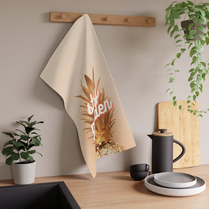 A beige kitchen towel with a graphic of a pineapple and the word 'stay golden' in pink script, hanging from a wooden peg on a kitchen wall above a countertop with dishes and plants.