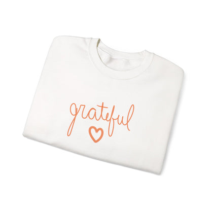 A folded white crewneck sweatshirt featuring the word 'grateful' in orange cursive script and an orange heart outline on the front, set against a plain white background.