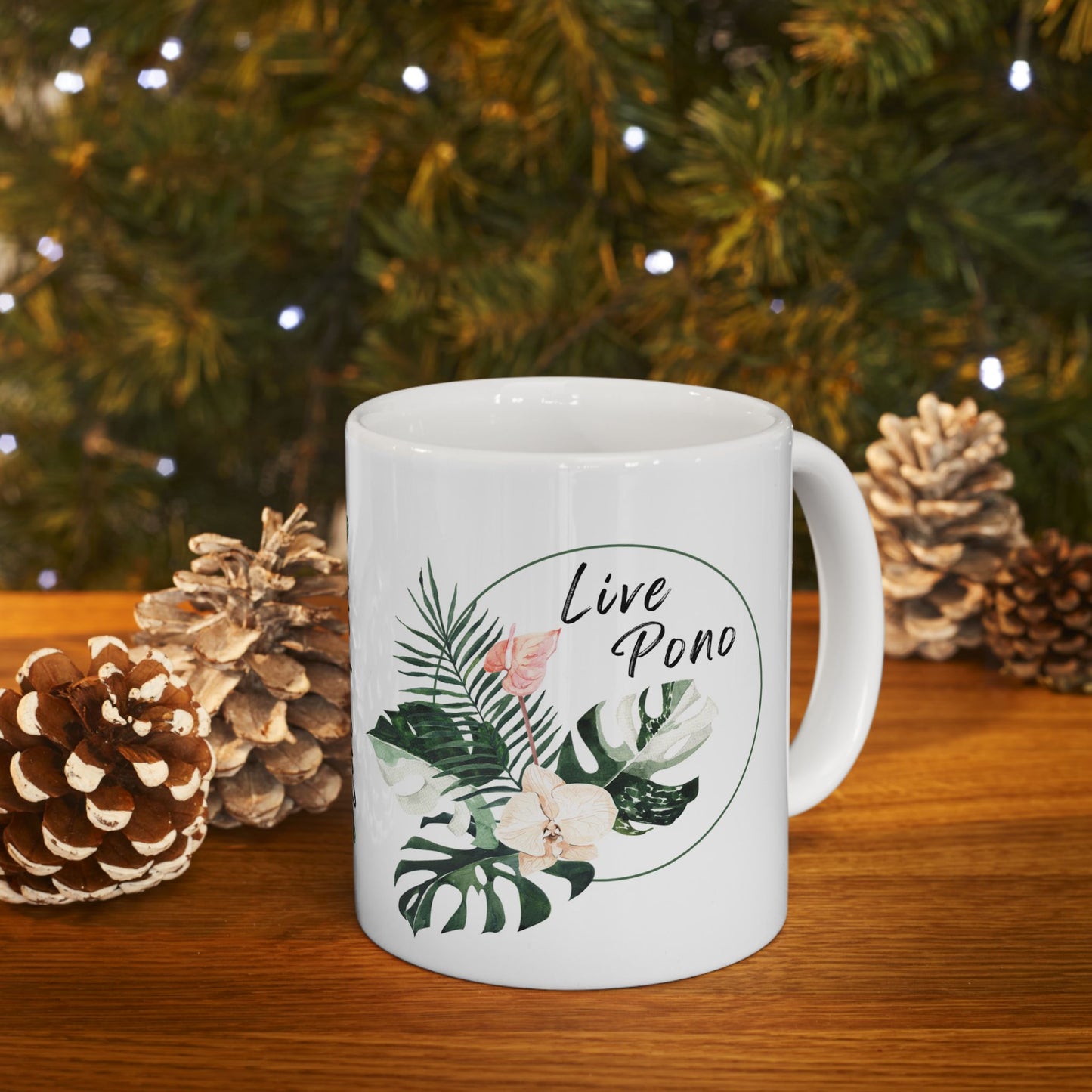 A white ceramic mug with a tropical leaf illustration and the words 'Live Pono' inside an oval, set on a wooden table with pinecones and a blurred Christmas tree background.