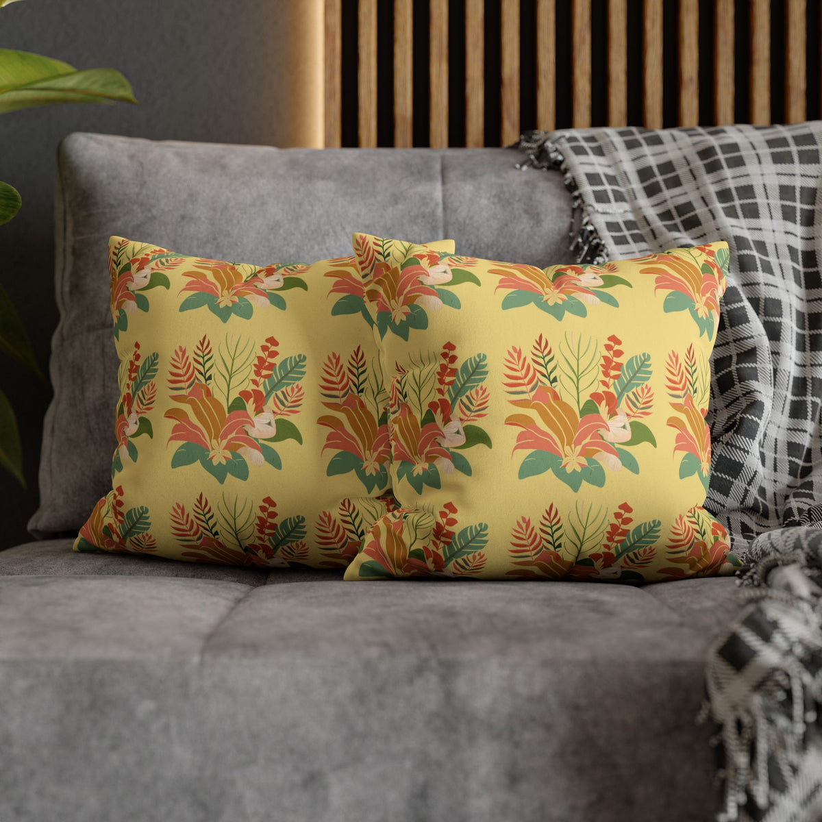 A rectangular throw pillow with a vibrant yellow background and a detailed tropical floral pattern in orange, green, and red, styled on a gray sofa next to a black and white plaid throw blanket.