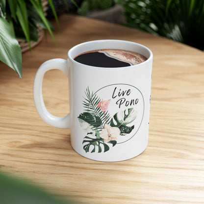 A white ceramic coffee mug filled with black coffee sits on a light wood table, decorated with tropical leaves and the words 'Live Pono' inside a circular frame. Green plants are visible in the softly blurred background.