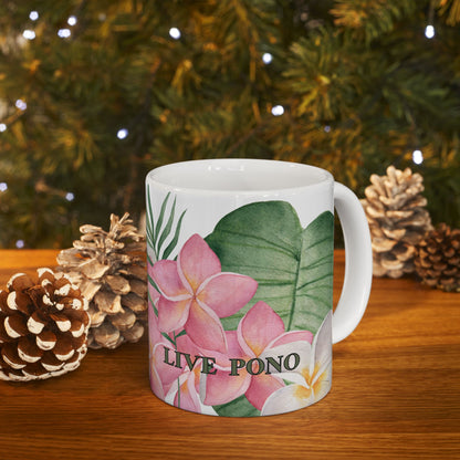 A white ceramic mug decorated with pink plumeria flowers, green tropical leaves, and the text 'LIVE PONO', set on a wooden table with pinecones and a blurred Christmas tree background.
