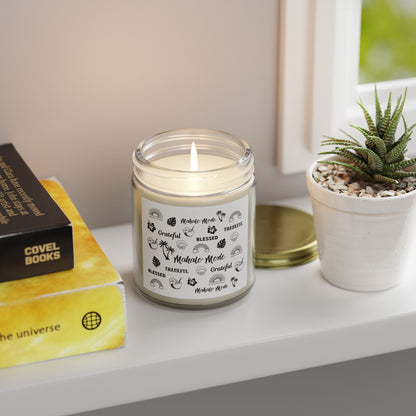 A glass jar candle with a gold metal lid and a white paper label featuring black tropical-themed illustrations and text like 'Mahalo Mode,' 'Grateful,' and 'Blessed,' shown on a white table with a plant and books.
