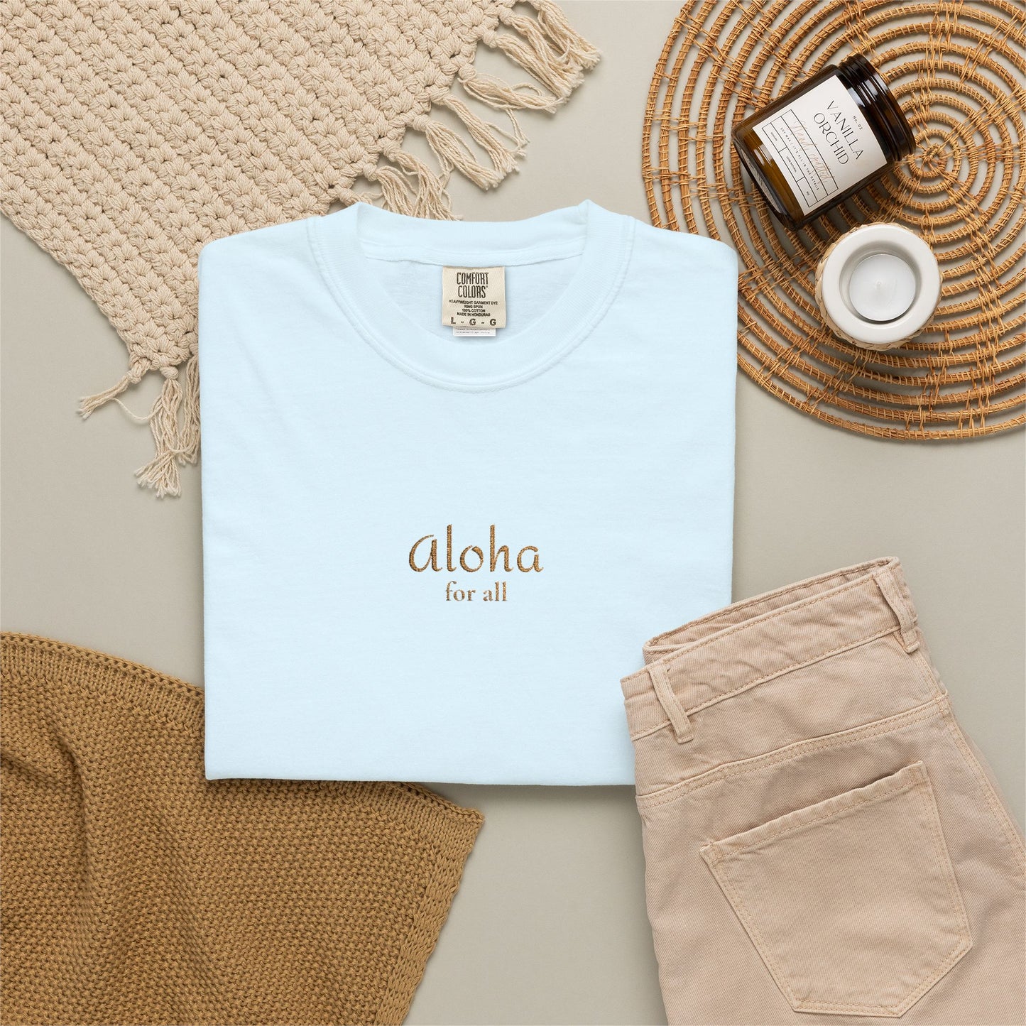 Light blue t-shirt with 'Aloha For All' text, beige pants, and decorative items on a neutral surface