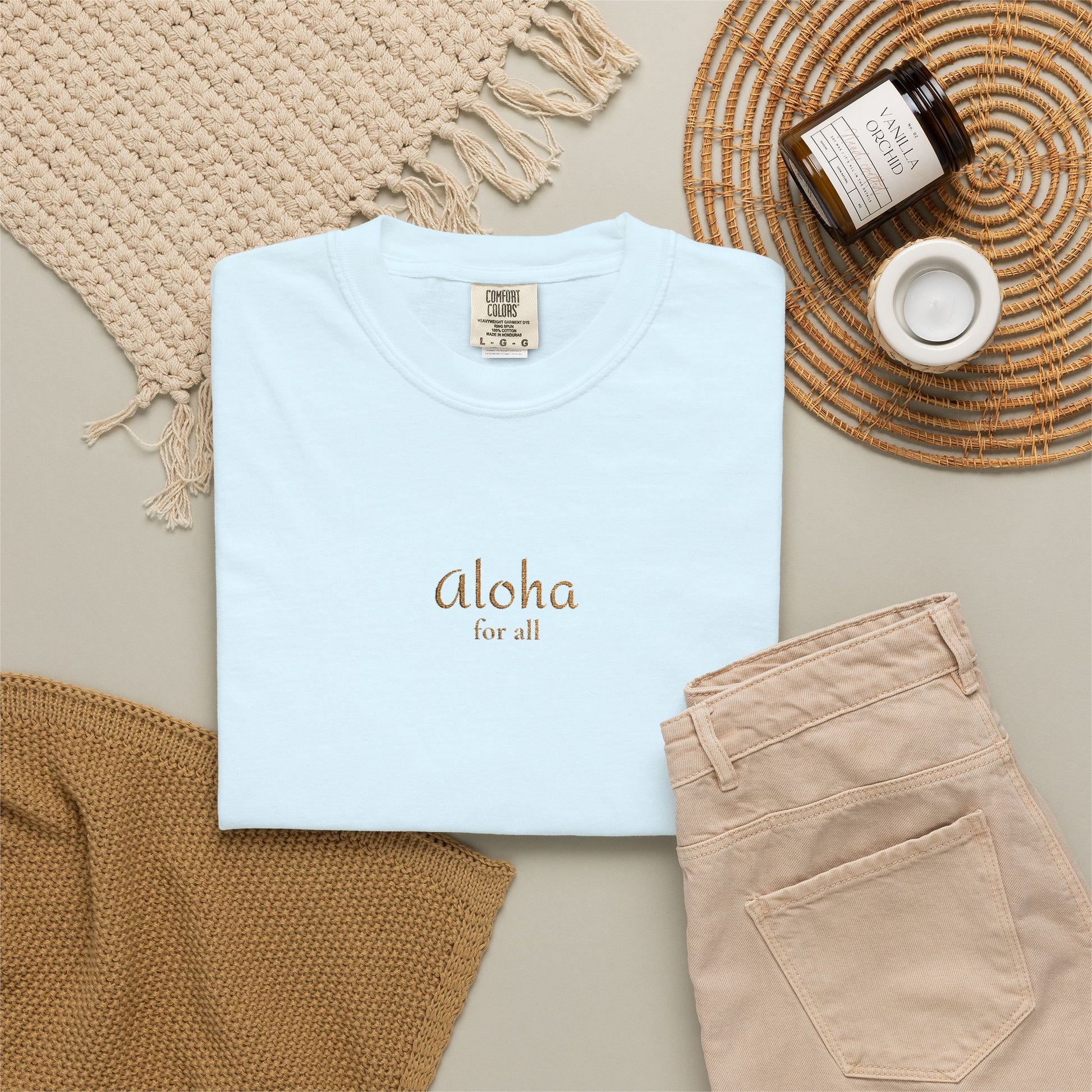 Light blue t-shirt with 'Aloha For All' text, beige pants, and decorative items on a neutral surface