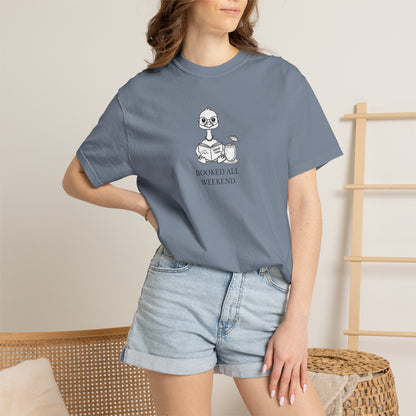 A woman standing wearing a blue comfort colors t-shirt with design of a nene goose reading a book and text 'booked all weekend' paired with denim shorts
