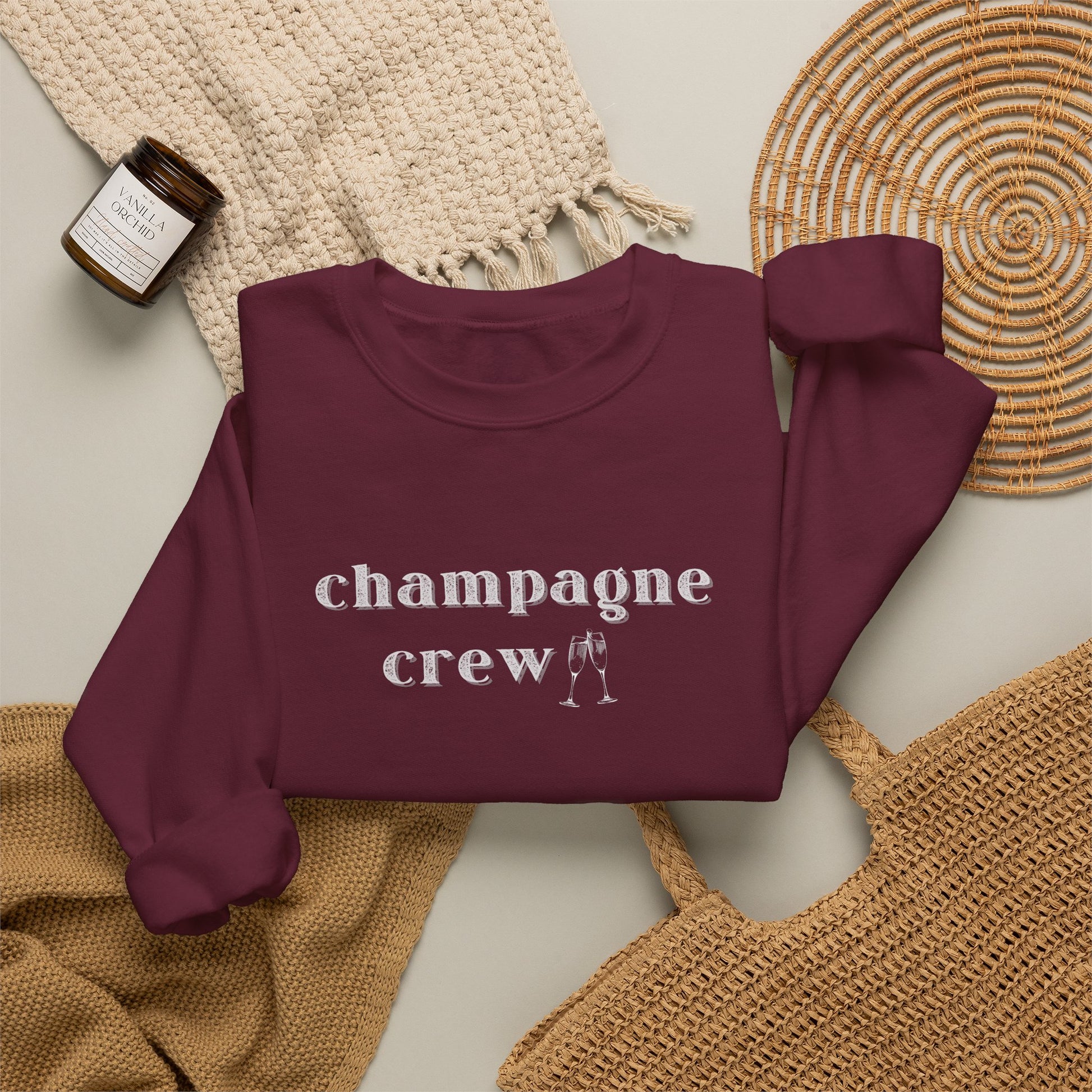 A maroon crewneck sweatshirt with the word 'champagne crew' printed in white lowercase serif letters across the chest, styled in a flat lay with a woven cream throw, a small amber candle jar, and a woven mat on a light beige background.
