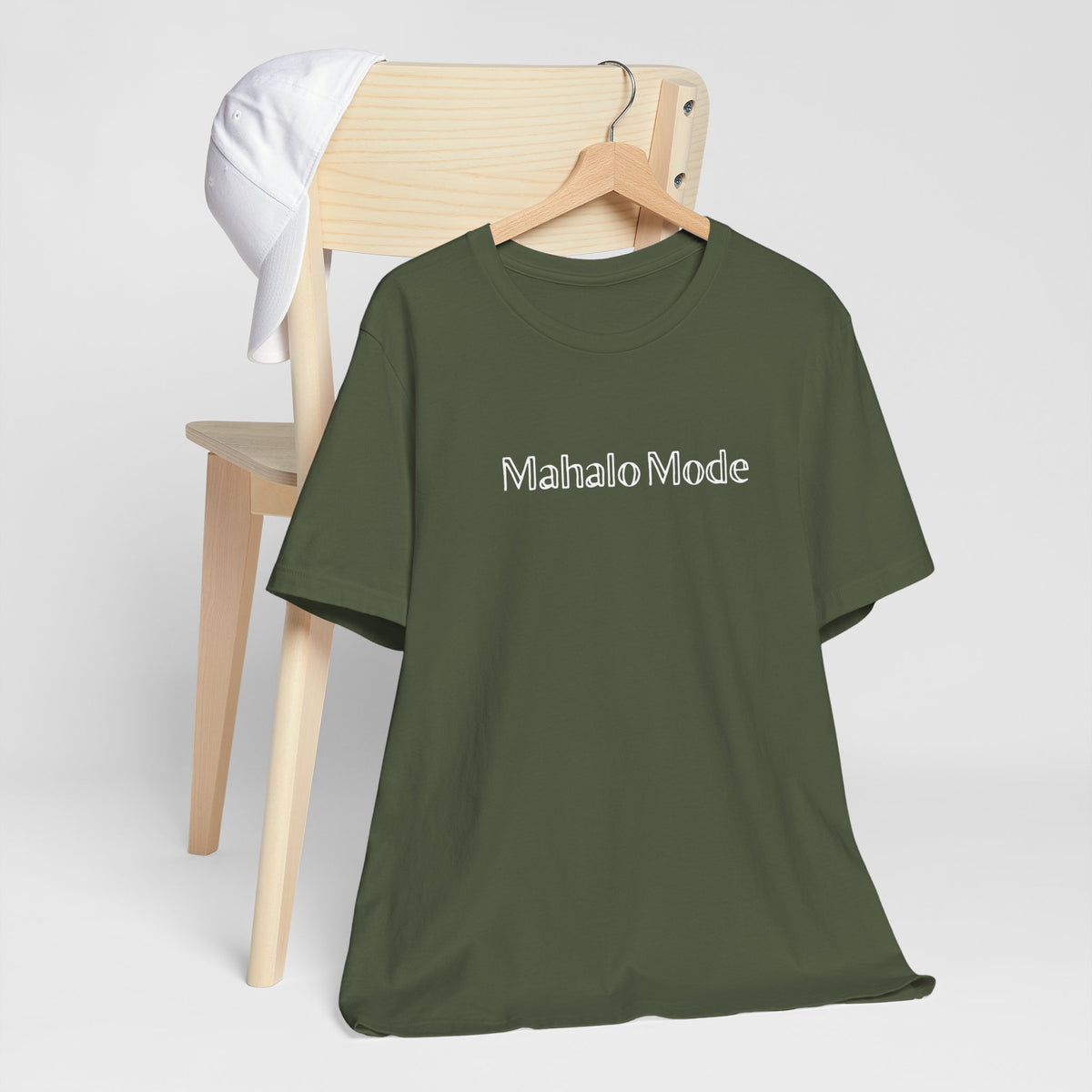 A Mahalo Mode graphic tee in olive green with white outlined lettering, Global Village Kailua original design, hanging from a wooden chair.