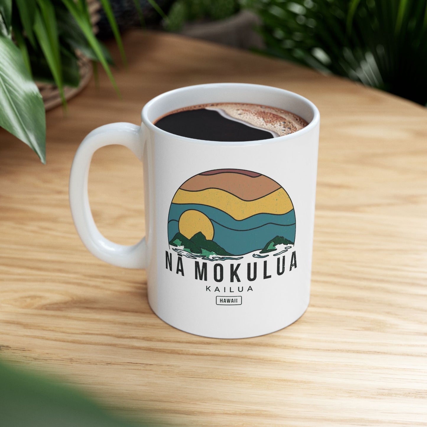 A white ceramic coffee mug with a colorful landscape graphic and the text 'NA MOKULUA KAILUA' on the front, filled with black coffee and placed on a light wood tabletop with blurred green plants in the background.
