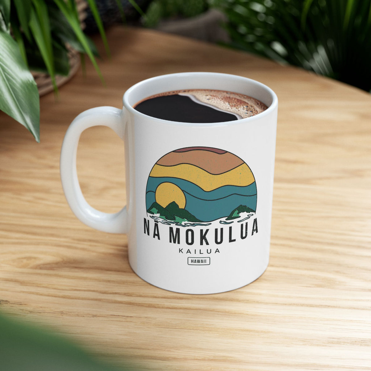 A white ceramic coffee mug with a colorful landscape graphic and the text 'NA MOKULUA KAILUA' on the front, filled with black coffee and placed on a light wood tabletop with blurred green plants in the background.