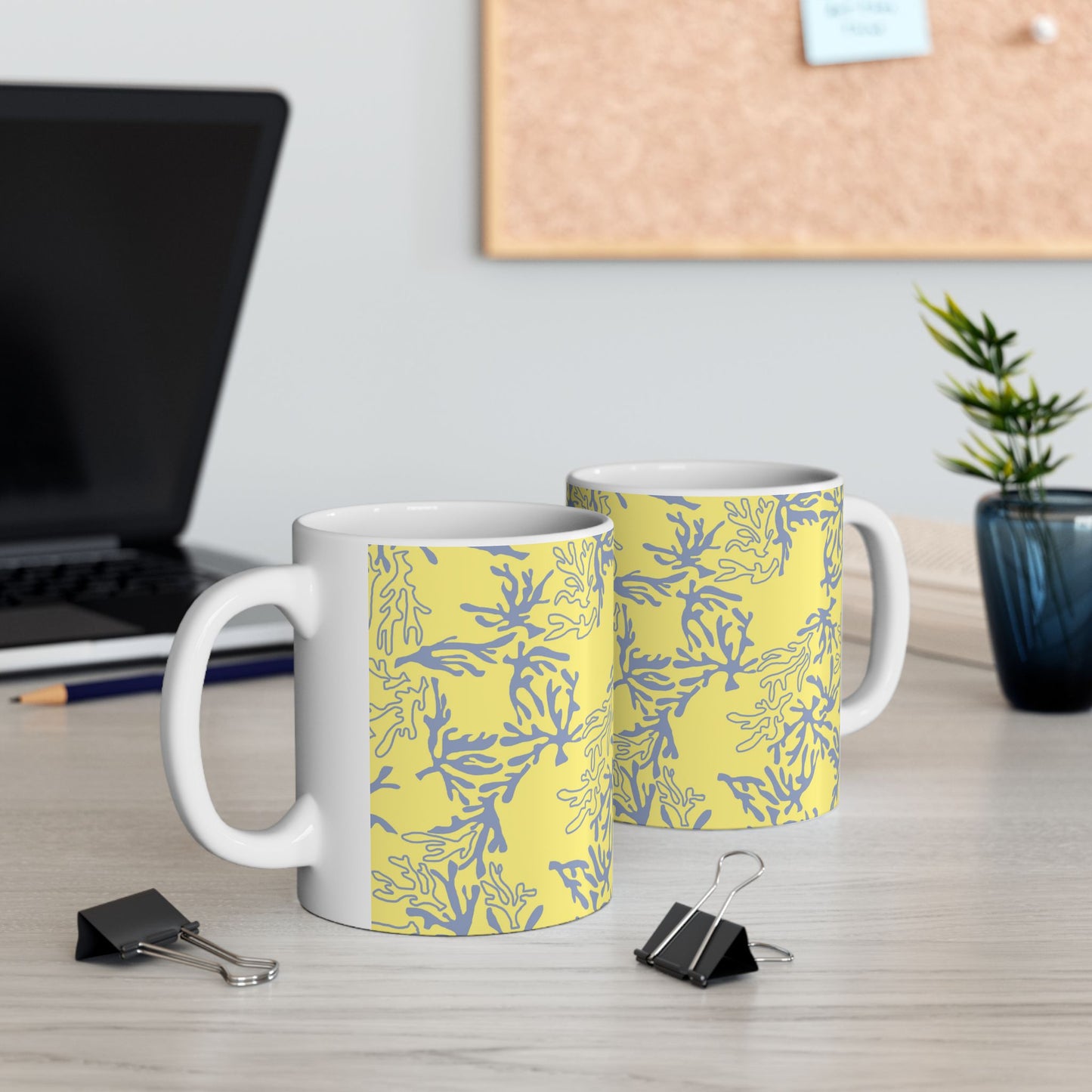 Two white ceramic mugs with a yellow and blue coral pattern, displayed on a light wood desk with office props like a laptop, binder clips, and a plant.
