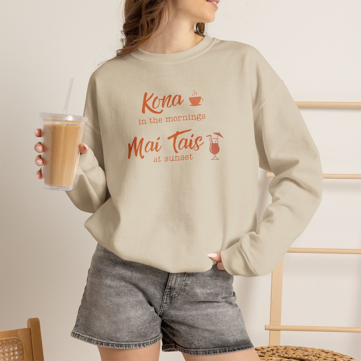 A woman holding an iced coffee wearing a beige crewneck sweatshirt featuring orange text that reads 'Kona in the mornings Mai Tais at sunset' with small coffee cup and cocktail glass illustrations.