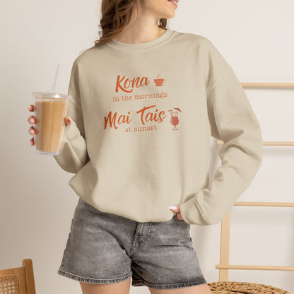A woman holding an iced coffee wearing a beige crewneck sweatshirt featuring orange text that reads 'Kona in the mornings Mai Tais at sunset' with small coffee cup and cocktail glass illustrations.
