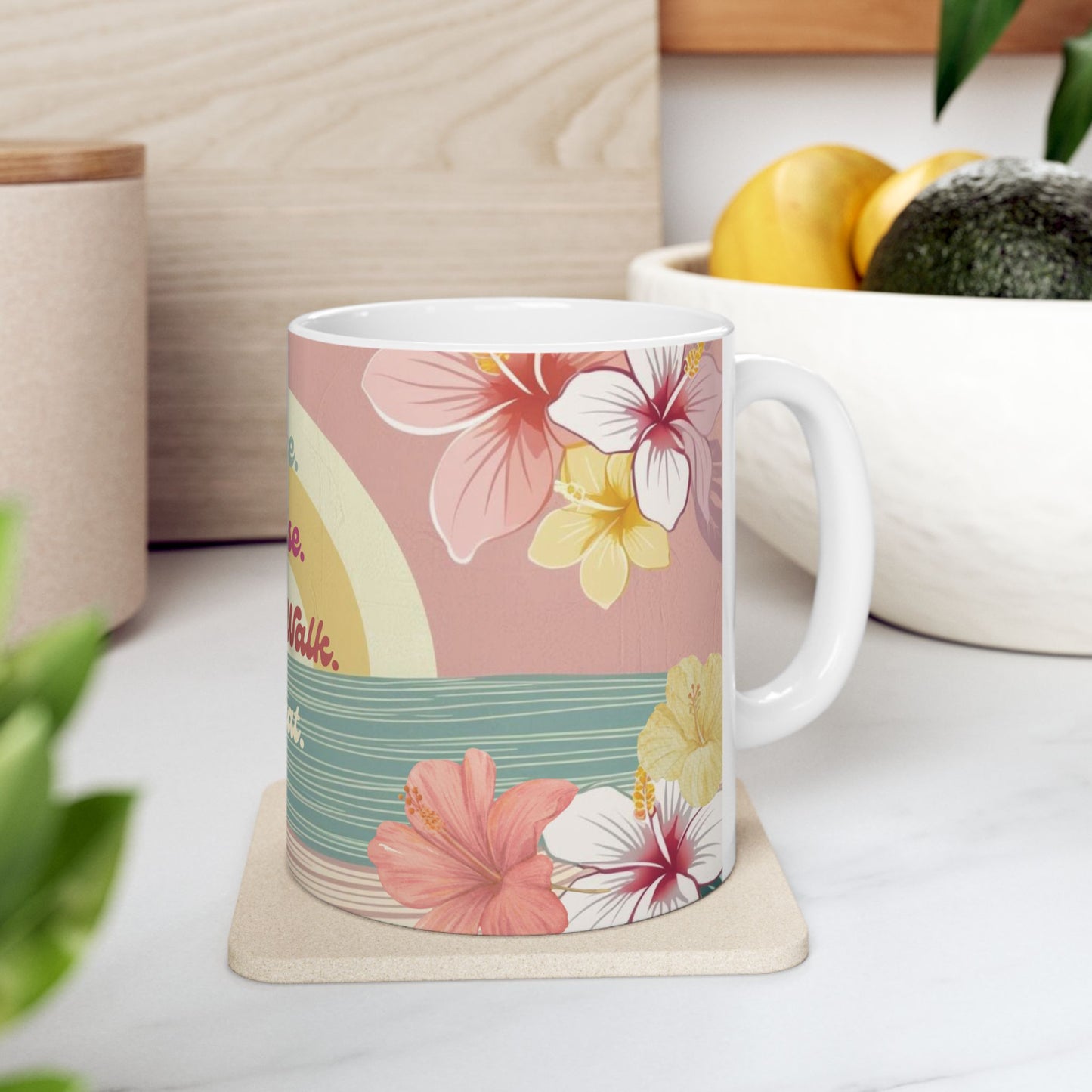 A white ceramic mug with a tropical floral print featuring pink, yellow, and white hibiscus flowers on a pink and teal background, resting on a beige coaster on a white marble countertop.