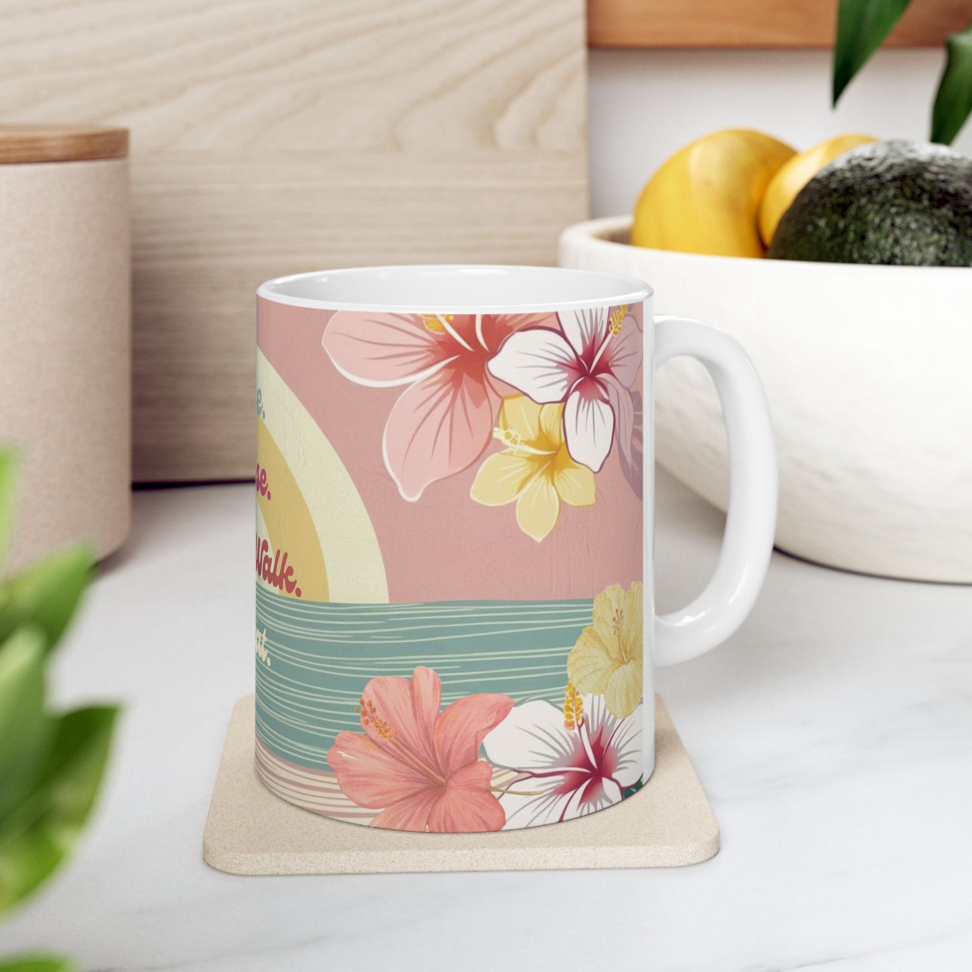 A white ceramic mug with a tropical floral print featuring pink, yellow, and white hibiscus flowers on a pink and teal background, resting on a beige coaster on a white marble countertop.